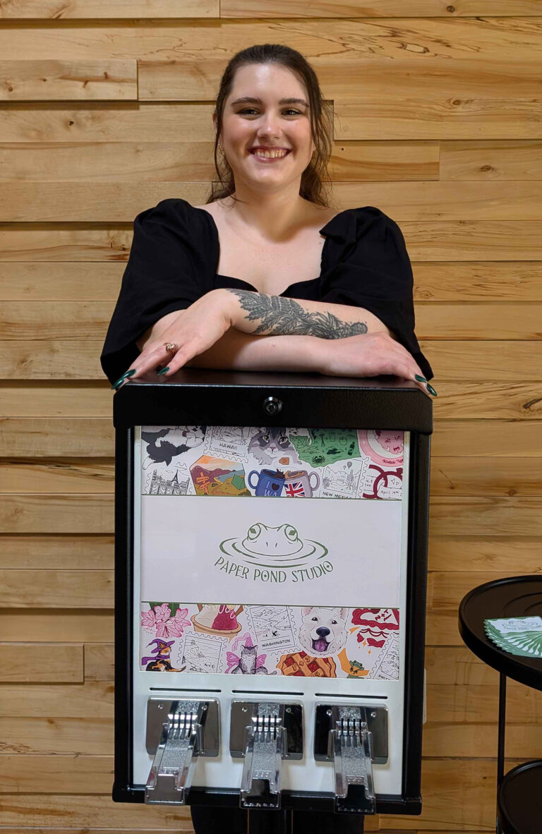 The owner of Paper Pond Studio leaning with crossed arms over the sticker machine displaying the Paper Pond Studio logo